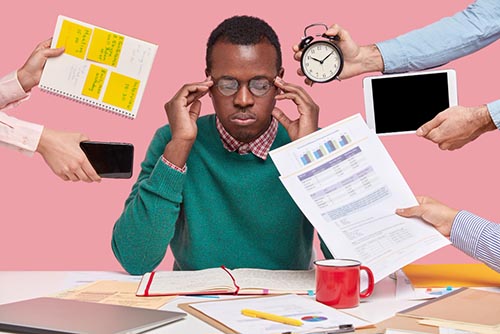 Worried black man sitting at a very messy desk holding his head as people are handing him more things to do.