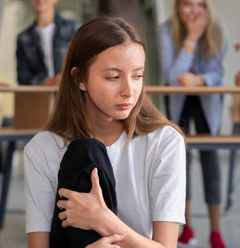 Socially anxious, worried young woman sitting alone with her leg up and arms wrapped around her leg while peers stare at her from afar