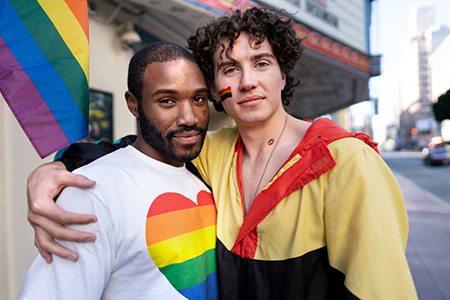 LGBTQ couple with arms around each other looking happy standing in front of pride flag.