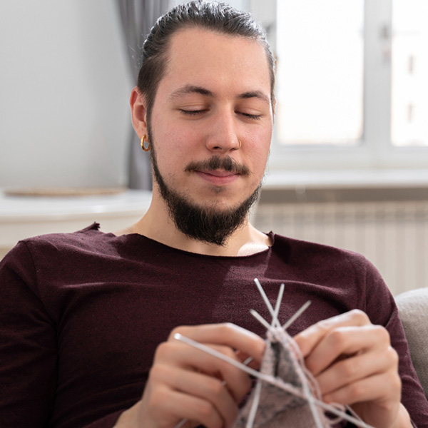 Neurodivergent male sitting on a couch knitting in the round with 5 needles.